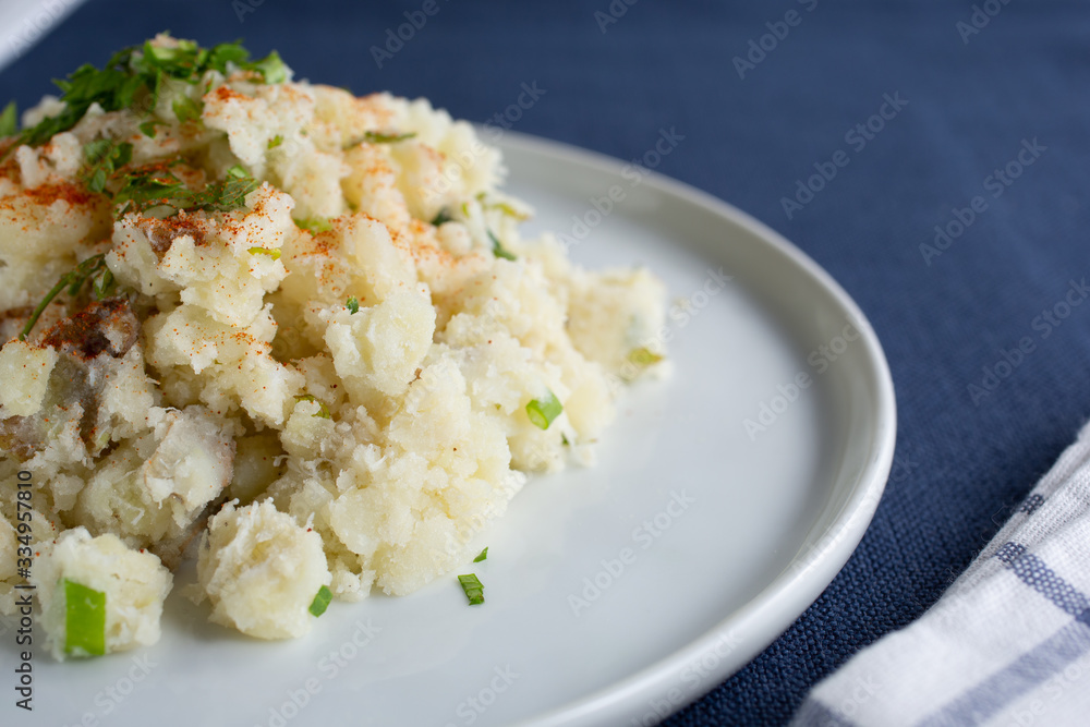 A closeup view of a plate of potato salad, cut off on the left side of the frame, in a restaurant or kitchen setting.
