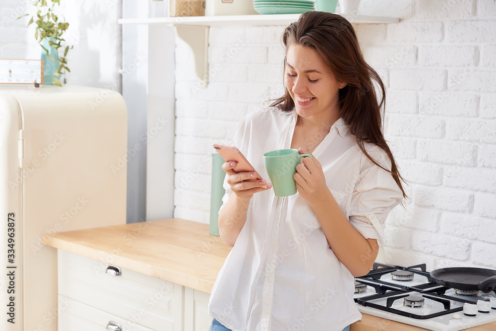 Young beautiful woman using cell phone and having a coffee in the kitchen Young beautiful woman using cell phone and having a coffee in the kitchen