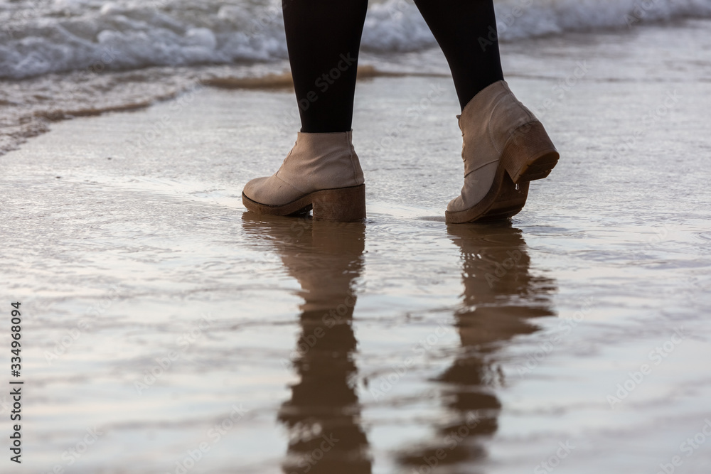 Girl's legs walking on the beach near water line in cream-colored winter shoes and black tights, water reflection 