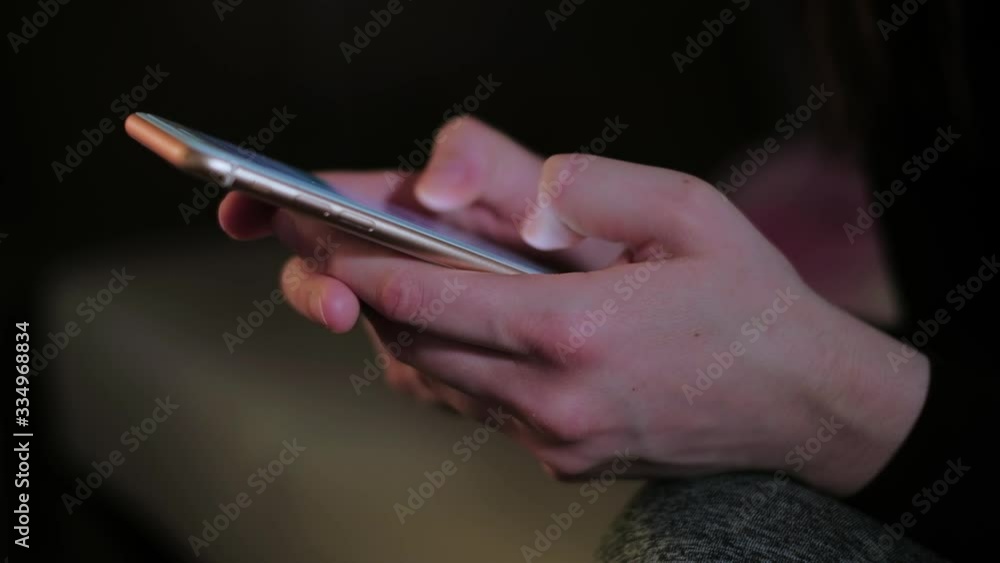 Young woman texting chatting on smart phone at night; girls finger tapping keypad, writing message on mobile devices touchscreen, natural low light; close up top side view Young woman texting chatting on smart phone at night; girls finger tapping keypad, writing message on mobile devices touchscreen, natural low light; close up top side view