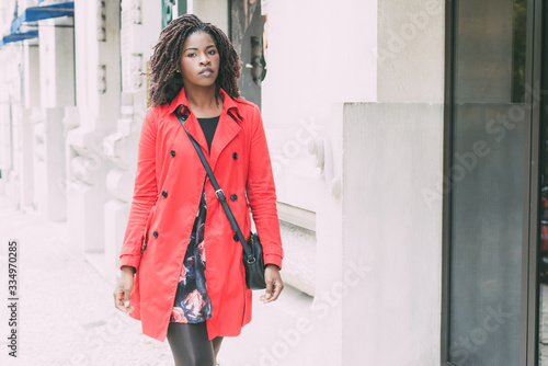 Attractive serious woman walking on street. Beautiful young woman in red trench coat walking in near building and looking at camera. Style concept