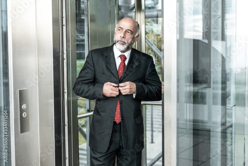 Serious confident mature businessman using office elevator. Elderly man in formal suit and tie standing in business canter glass interior. Business portrait concept