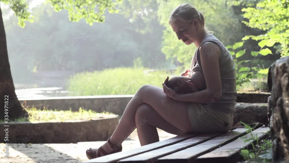 A girl sitting on a bench feeds the child and strokes the head, sharply raises her eyes and looks towards the camera and smiles. Long shot. Full HD, no sound. A girl sitting on a bench feeds the child and strokes the head, sharply raises her eyes and looks towards the camera and smiles. Long shot. Full HD, no sound.
