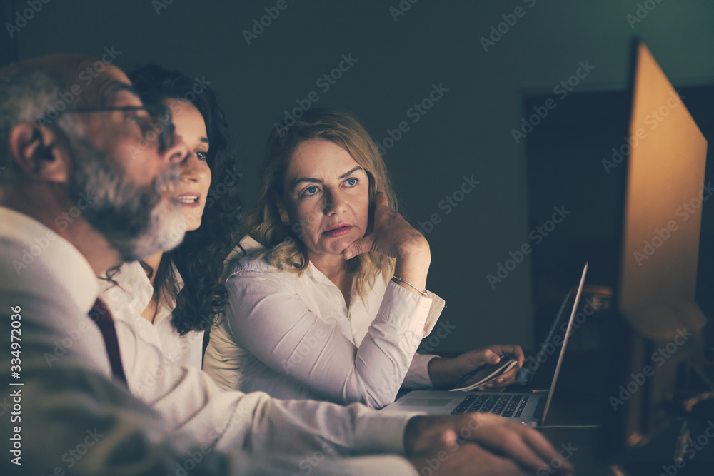 Male and female coworkers discussing project in dark office. Side view of focused business people sitting at workplace and using desktop computer in dark office. Working late concept Male and female coworkers discussing project in dark office. Side view of focused business people sitting at workplace and using desktop computer in dark office. Working late concept