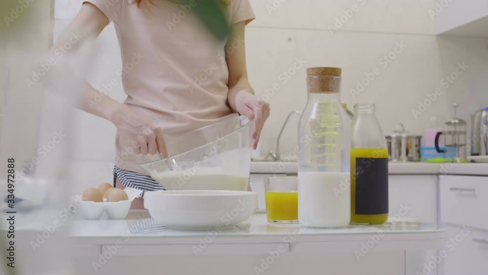Sequence of shots of unrecognizable woman standing at worktop in the kitchen and mixing milk, eggs, flour and sunflower oil in glass bowl Sequence of shots of unrecognizable woman standing at worktop in the kitchen and mixing milk, eggs, flour and sunflower oil in glass bowl
