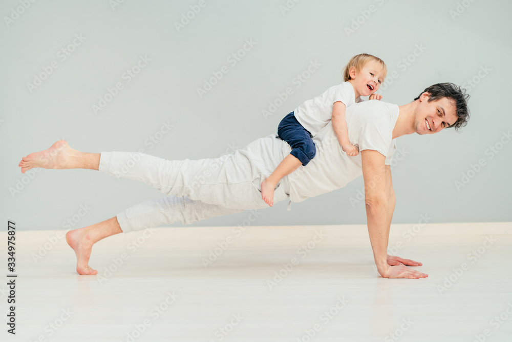 Positive sportive father doing pilates, yoga exercise, workout at home in floor with his little baby son having fun, excited emotion, play together, communication concept.