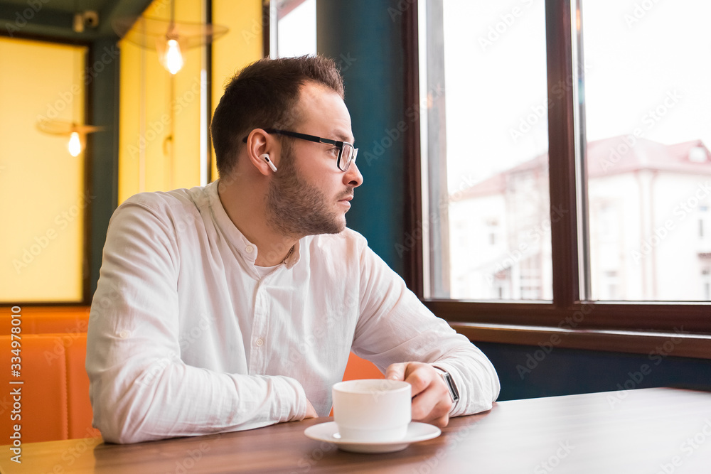 Young attractive businessman in a white shirt, glasses and headphones drinks coffee and looks thoughtfully out the window Young attractive businessman in a white shirt, glasses and headphones drinks coffee and looks thoughtfully out the window