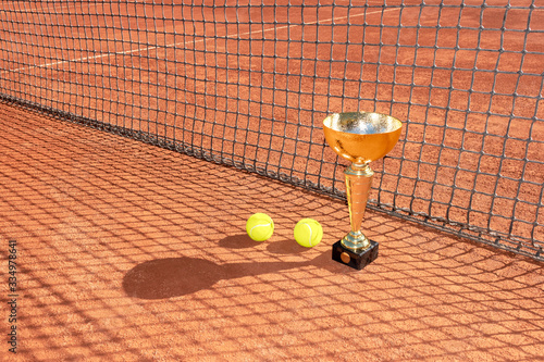 Trophy cup and tennis balls on a red clay court and shadow from the net. Champion or victory trophy. Winner in tennis tournament concept. Sports background. Copy space photo