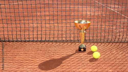 Golden champion trophy and balls on a red clay tennis court and net shadow. Winner in tennis tournament concept. Sports background. Copy space © Elena