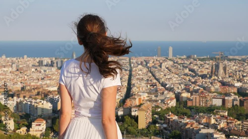 Woman stay on height against cityscape, view from behind, turn head, quickly look aside and turn back to urban landscape. Lady rest at windy top of hill, overlook to Barcelona photo