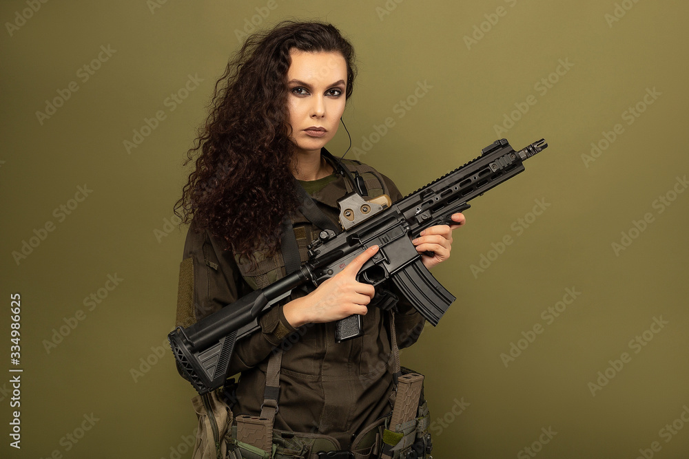 Military woman in green uniform with a vest unloading with a machine gun in her hands on a khaki background. Military woman in green uniform with a vest unloading with a machine gun in her hands on a khaki background.