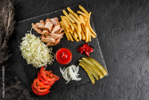 French fries potatoes, grilled chicken meat, pickles and cabbage, tomatoes and ketchup sauce on black stone background. Hot fast food dish, top view
