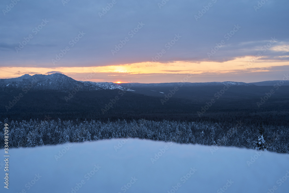 Winter forest with snow-covered fir trees high in the mountains. Dawn with bright colors on the horizon far away in the mountains. Golden clouds with the first rays of the sun.