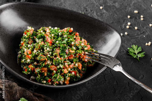 Tabbouleh salad with bulgur, tomatoes, parsley, green onion, bell pepper in plate on black stone background. Traditional middle eastern or arabic dish. Healthy food, vegetarian dieting, close up photo