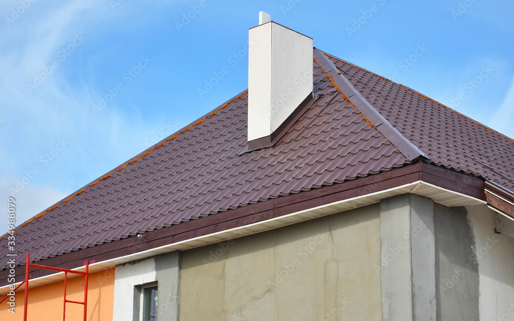 Close-up on a dark red metal tiled bonnet roof with a white chimney and stucco finish walls with scaffolding.