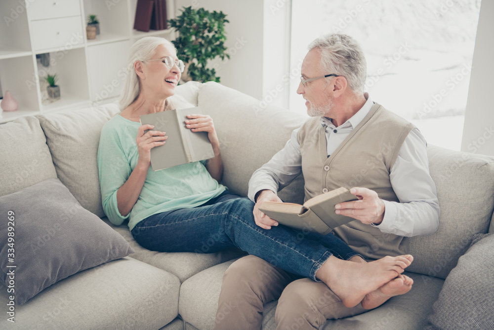 Portrait of cheerful grandmother grandfather hold fiction book laugh wear eyeglasses eyewear denim jeans trousers teal brown pullover sit lie divan bare foot in room Portrait of cheerful grandmother grandfather hold fiction book laugh wear eyeglasses eyewear denim jeans trousers teal brown pullover sit lie divan bare foot in room