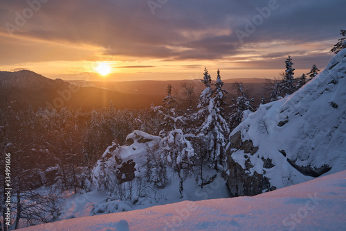 Winter forest with snow-covered fir trees high in the mountains. Dawn with bright colors on the horizon far away in the mountains. Golden clouds with the first rays of the sun. © nikolay_alekhin