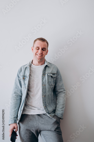 a young man in a white t shirt and denim jacket stands against a white wall