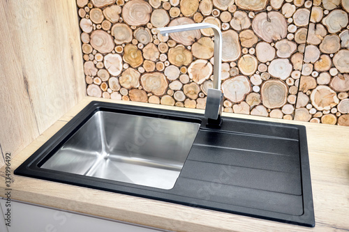 Modern design chrome water tap over stainless steel kitchen sink on table top made of natural wood with surface behind countertop decorated with wall cross section of tree trunks cut tree logs photo