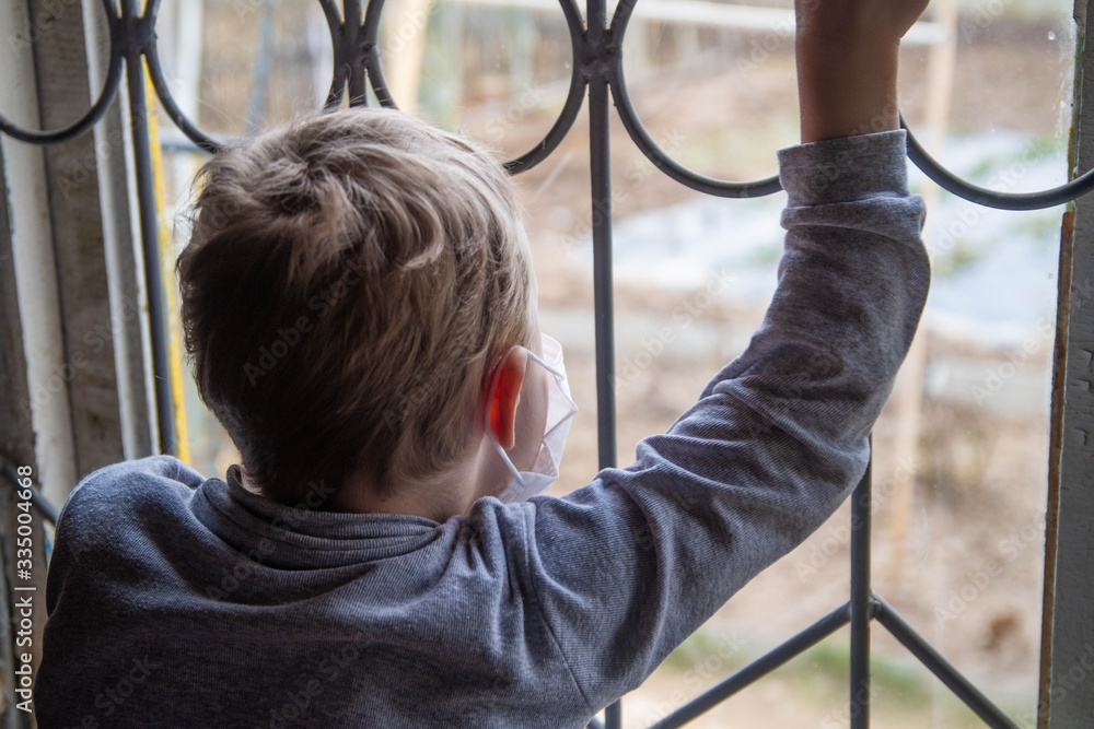 Obraz premium Boy kid blond European sitting in a medical mask in front of the window of the house. Isolation, quarantine. Virus epidemic and pandemic.