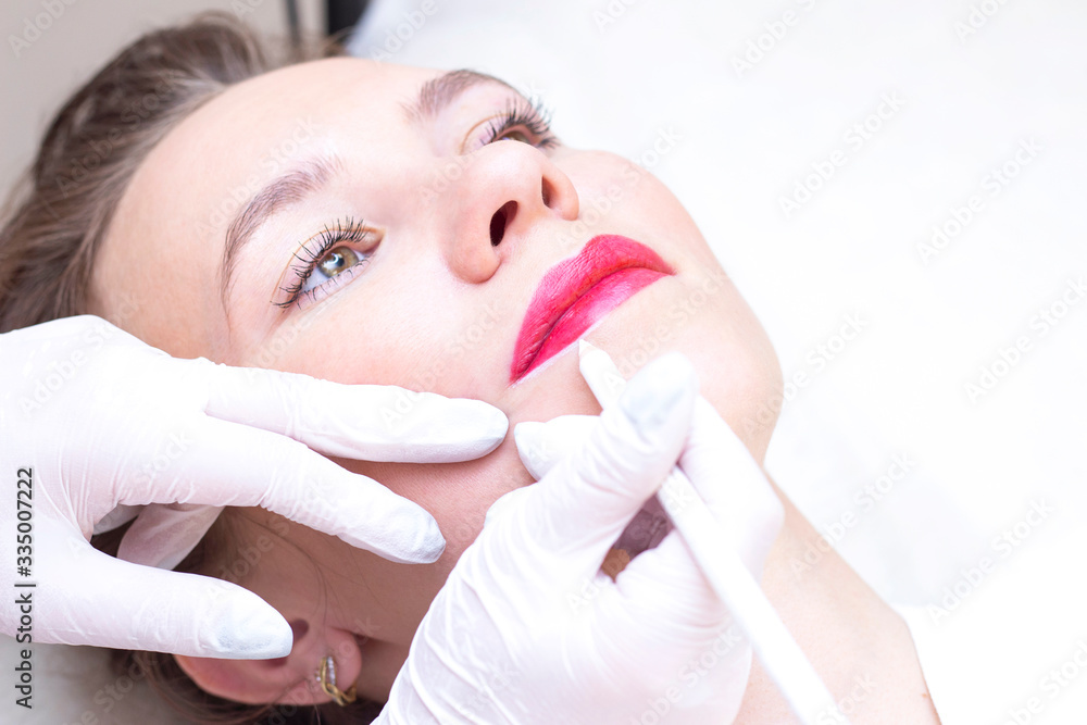 Young woman having permanent makeup on her lips at the beauticians salon. Permanent Makeup (Tattoo). drawing a contour with a white lip pencil Young woman having permanent makeup on her lips at the beauticians salon. Permanent Makeup (Tattoo). drawing a contour with a white lip pencil