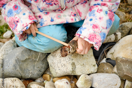 Closeup of low section of 4 year old  caucasian child girl in warm clothing sitting on stones in the garden and playing with two wooden sticks. Seen in March in Germany.