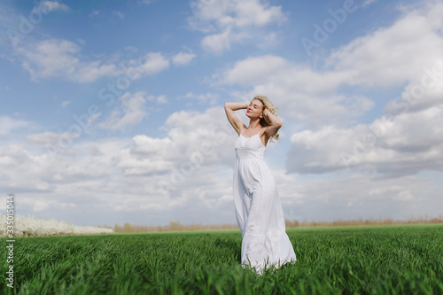  blonde girl in a white dress sits on the green fresh spring grass, blue sky
