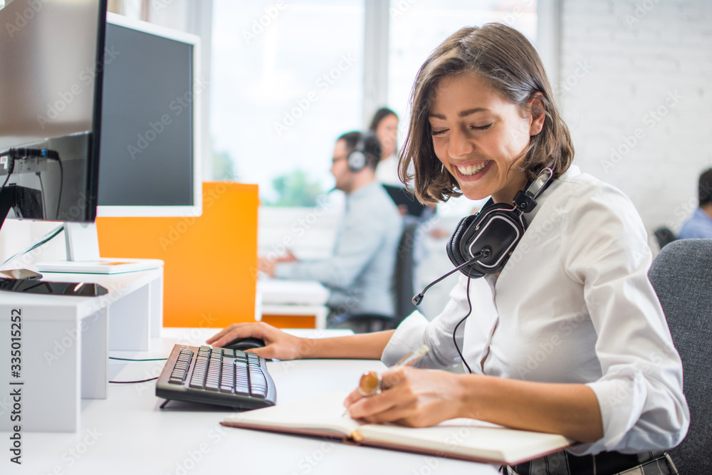 Young businesswoman working in front of computer, writing notes to notebook while sitting at her workplace in office Young businesswoman working in front of computer, writing notes to notebook while sitting at her workplace in office