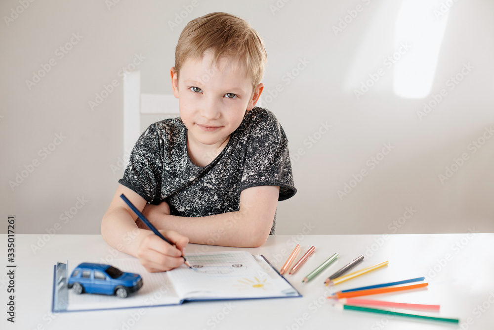 A child of European appearance is sitting at a table drawing with pencils in a white interior A child of European appearance is sitting at a table drawing with pencils in a white interior