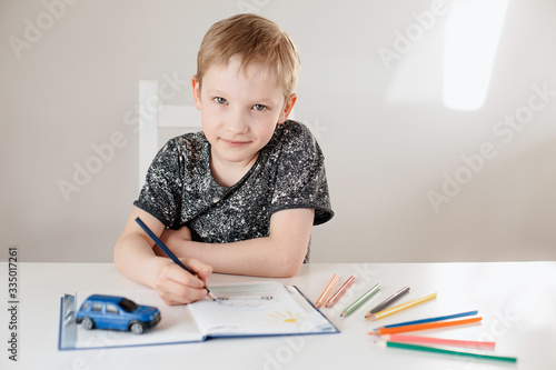 A child of European appearance is sitting at a table drawing with pencils in a white interior