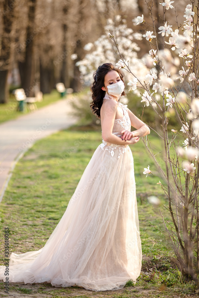 Beautiful young bride in a wedding dress and a white medical mask on her face near a blooming magnolia.  Kovid-19 protects.