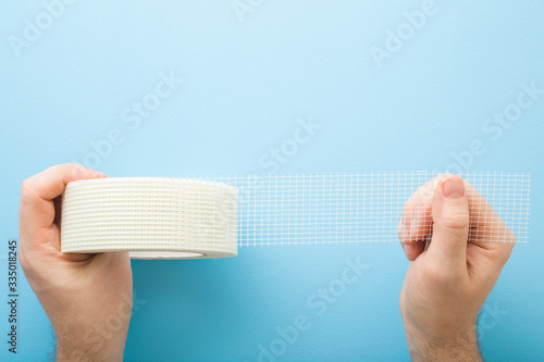 Young man hands stretching roll of mesh for split or seam on wall or ceiling. Light pastel blue table background. Closeup. Point of view shot. Top down view.