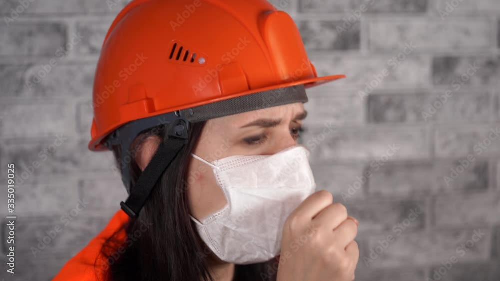 Female construction worker in overalls and medical mask coughing on background of gray brick wall. Concept of threat of infection. Female construction worker in overalls and medical mask coughing on background of gray brick wall. Concept of threat of infection.