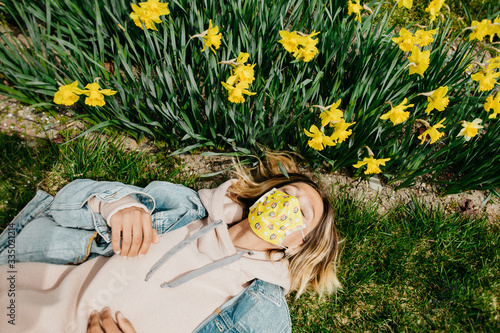 Beautiful young woman in the daffodils field wearing yellow medical mask, lying on the green grass. Spring, sunny day, natural light