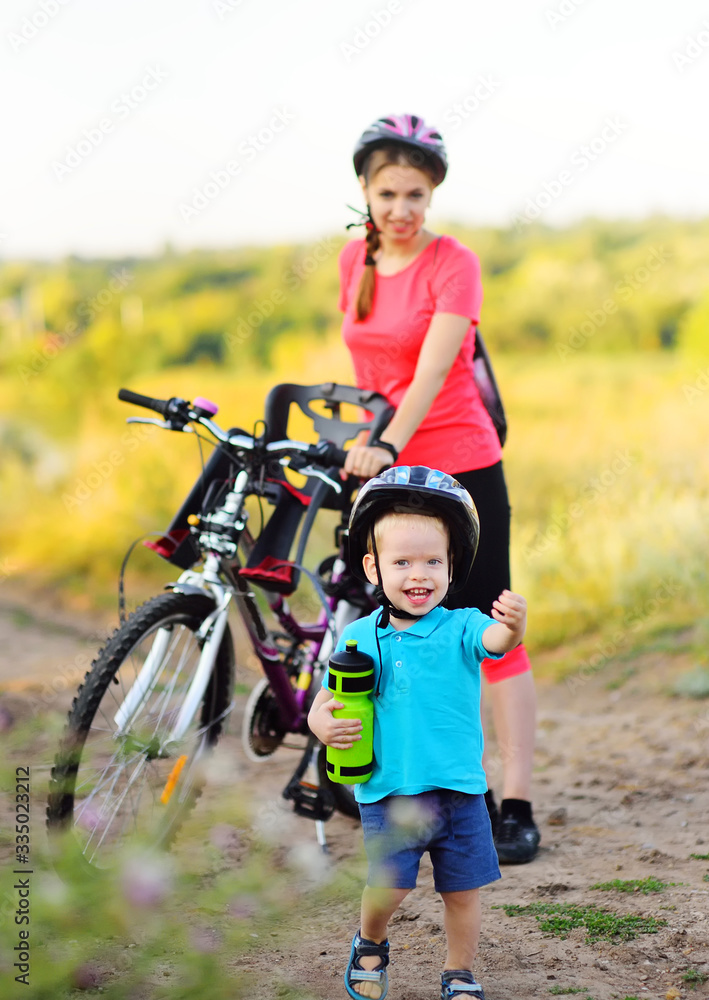 a small happy baby boy in a protective Bicycle helmet and with a bottle of water in his hands is smiling against the background of his mother on a Bicycle and nature.