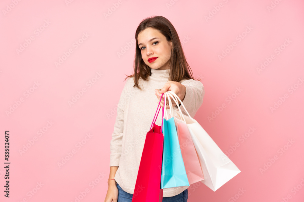 Teenager girl isolated on pink background holding shopping bags and giving them to someone