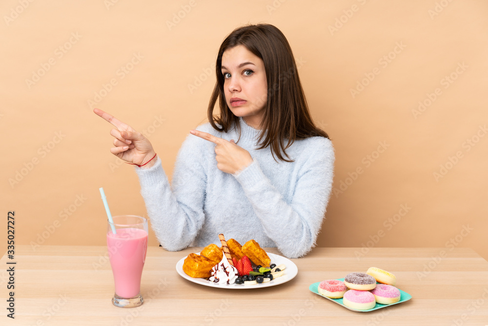 Teenager girl eating waffles isolated on beige background frightened and pointing to the side Teenager girl eating waffles isolated on beige background frightened and pointing to the side