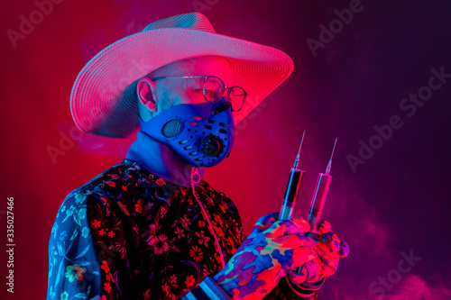 Strange man in medical respirator mask, glasses and hat holding two syrenges with coronavirus covid 19 test on red blue background.with smoke. Evil doctor, inventor of corona virus covid 19 conept photo