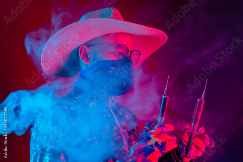 Strange man in medical respirator mask, glasses and hat holding two syrenges with coronavirus covid 19 test on red blue background.with smoke. Evil doctor, inventor of corona virus covid 19 conept photo