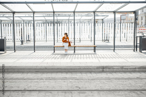 Woman sitting alone at the public transport stop on a sunny day outdoors, general plan on a modern stop. Concept of a transportation and urban life
