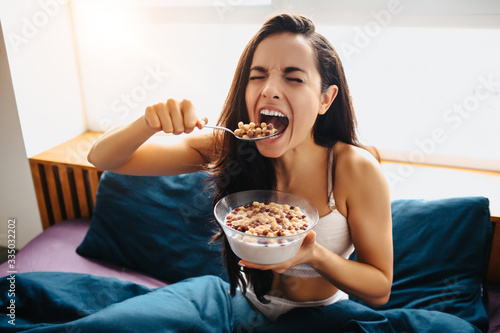 Young beautiful woman in morning bed at home. Hungry female person divour meal or food. Healthy breakfast cereal with milk in bowl. photo