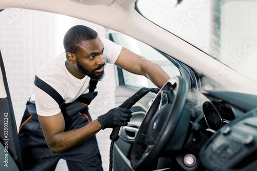 Auto cleaning service and detailing concept. Handsome African man in uniform cleaning interior of the car with hot steam cleaner. Selective focus.