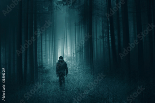 Man walking alone in magical dark turquoise green colored foggy artistic forest landscape.