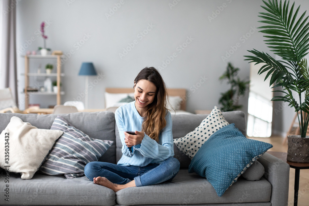 Online chat. Smiling woman talking on smartphone with friends while sitting on sofa at home. - obrazy, fototapety, plakaty 