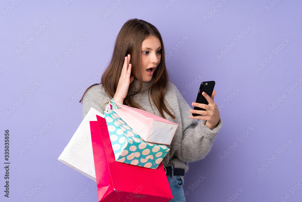 Teenager caucasian girl isolated on purple background holding shopping bags and writing a message with her cell phone to a friend Teenager caucasian girl isolated on purple background holding shopping bags and writing a message with her cell phone to a friend