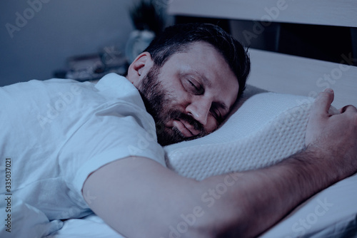 A lonely bearded dark-haired man sleeps soundly on his stomach at night on the bed. Close-up, orthopedic pillow, bedroom, self-isolation.
