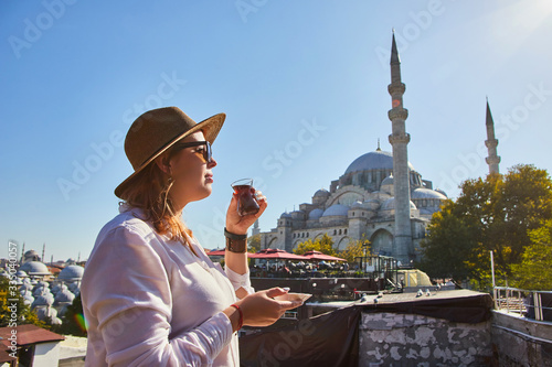 Photography Happy attractive woman tourist in hat, drinks traditional turkish tea (armudas) against the background of Suleymaniye mosque, Istanbul, Turkey