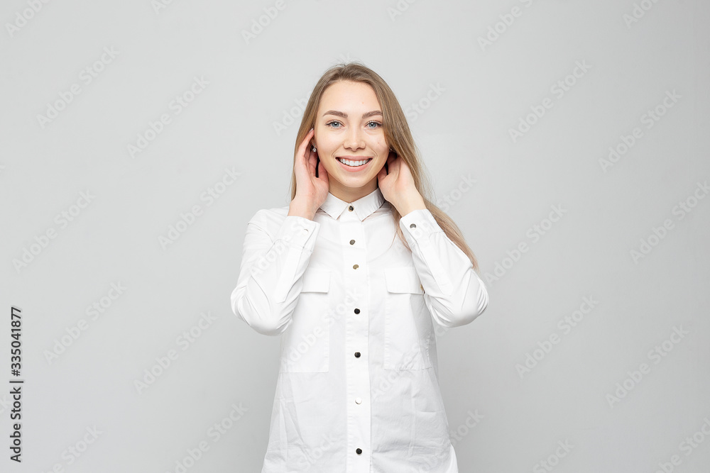 Beautiful young woman portrait. Portrait of a beautiful smiling blonde in white shirt on a white-gray. woman straightens her hair or holds her head Beautiful young woman portrait. Portrait of a beautiful smiling blonde in white shirt on a white-gray. woman straightens her hair or holds her head