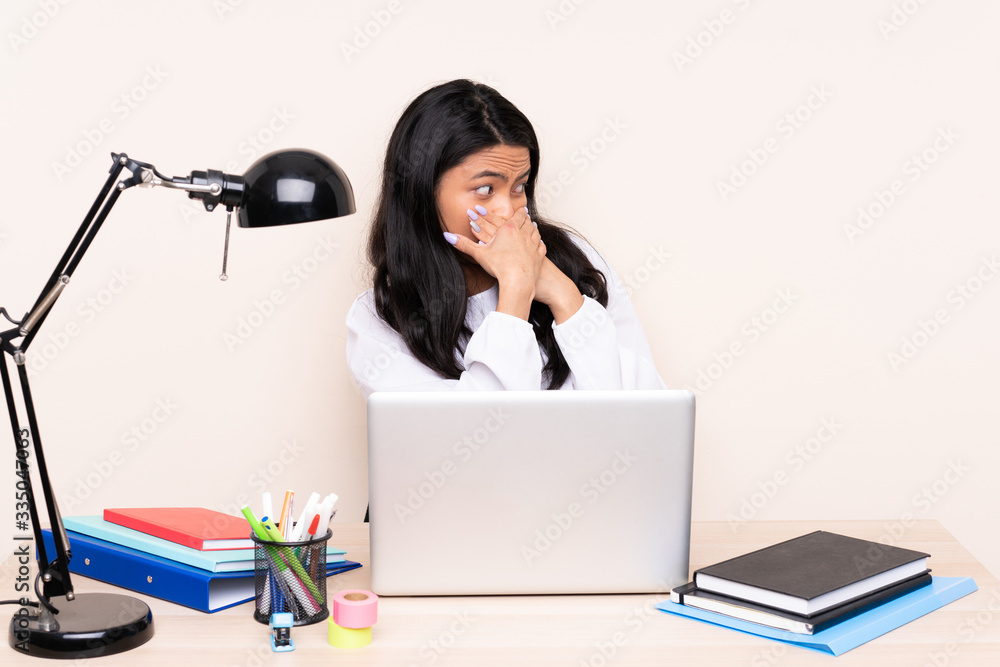Student asian girl in a workplace with a laptop isolated on beige background covering mouth and looking to the side Student asian girl in a workplace with a laptop isolated on beige background covering mouth and looking to the side