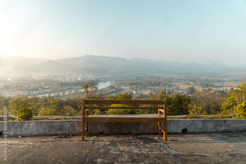 A beautiful photo of an empty bench on top of a mountain overlooking a sunset and a small town with the mountains in the distance